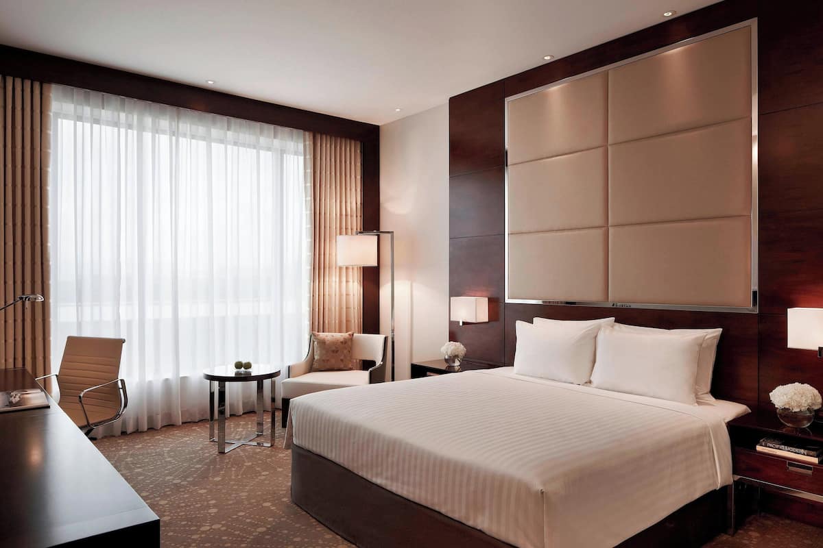 A modern hotel room with a large bed, a desk, a chair, a small table, and a large window with curtains.