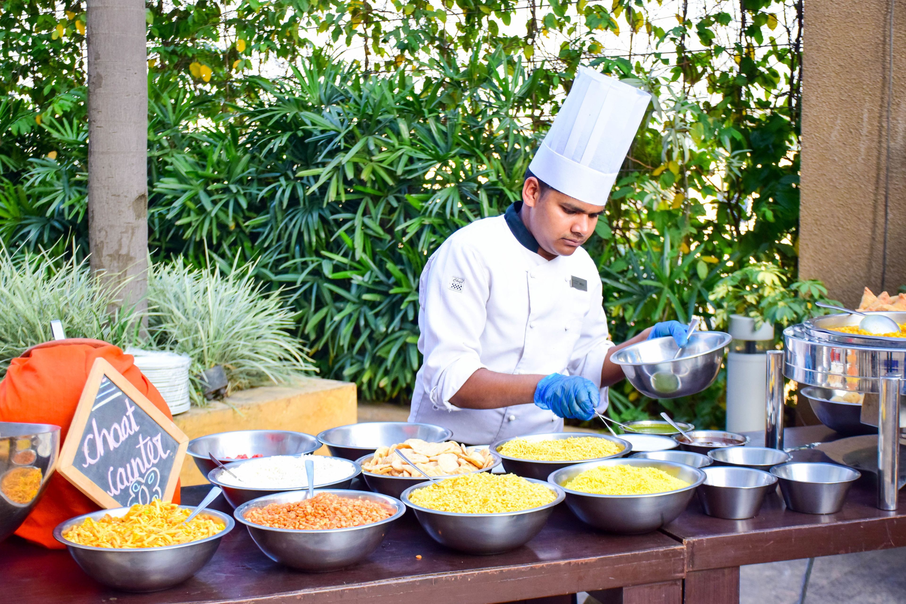 A buffet setup with various food items in metal bowls, a chef in uniform, and a sign that reads "chot launtr".