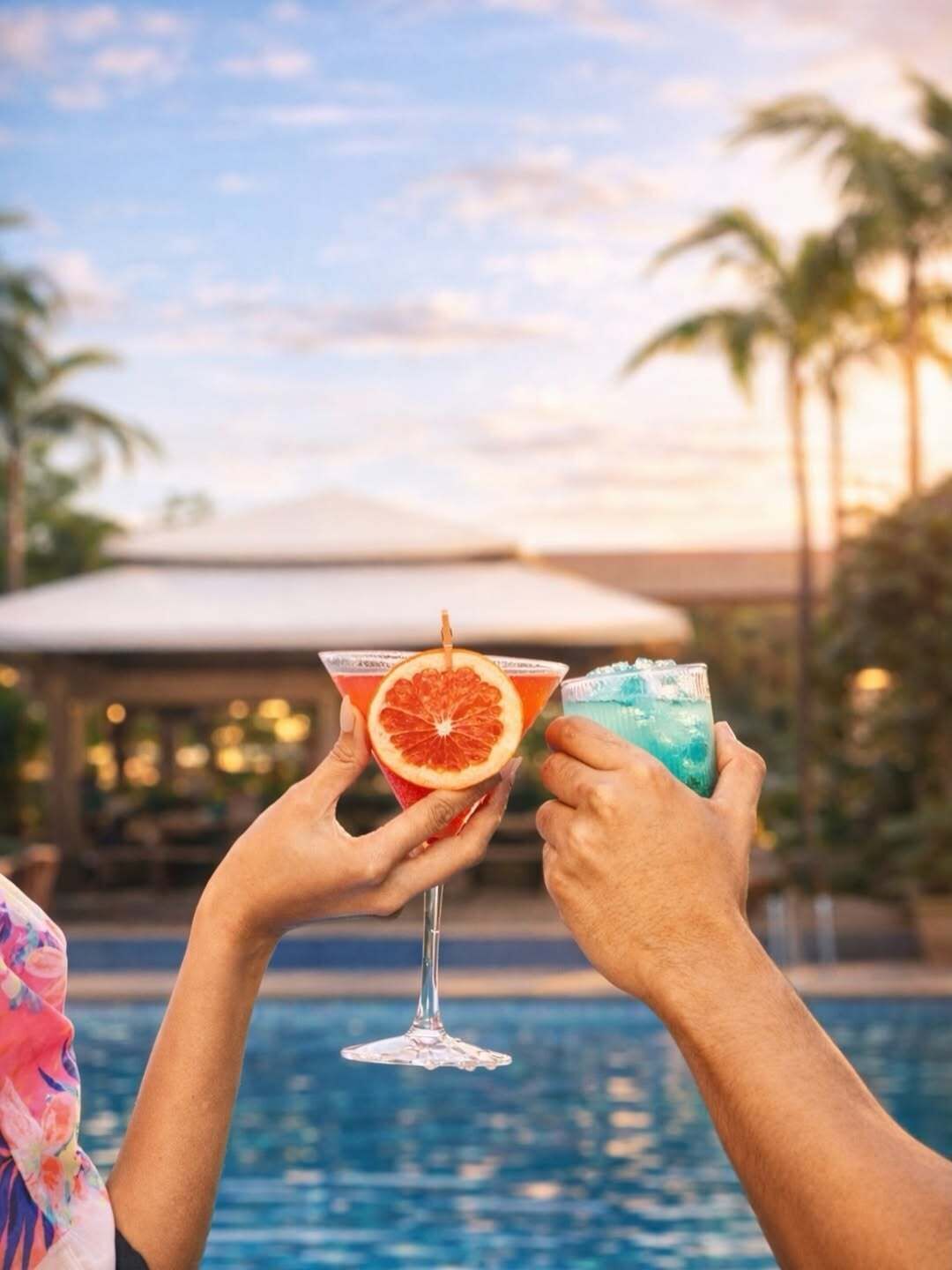 Two people holding cocktail glasses with a pool and palm trees in the background.