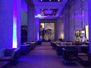 A lobby with a modern design, featuring a central seating area with sofas and armchairs, a large potted plant, and a glass entrance leading to a bar area. - Courtyard by Marriott Pune Chakan (Rajgurunagar)