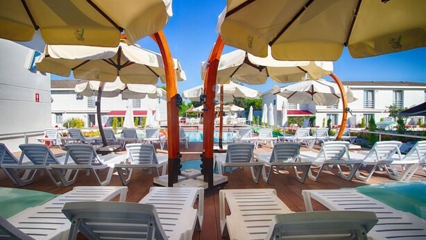 A poolside area with white lounge chairs and beige umbrellas, surrounded by a wooden deck and a pool. - Vela Apart Hotel (Cesme)