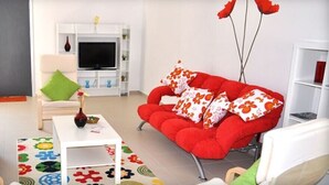 A living room with a white sofa, a red sofa with floral cushions, a white coffee table, and a TV stand with a flat-screen TV. - Vela Apart Hotel (Cesme)