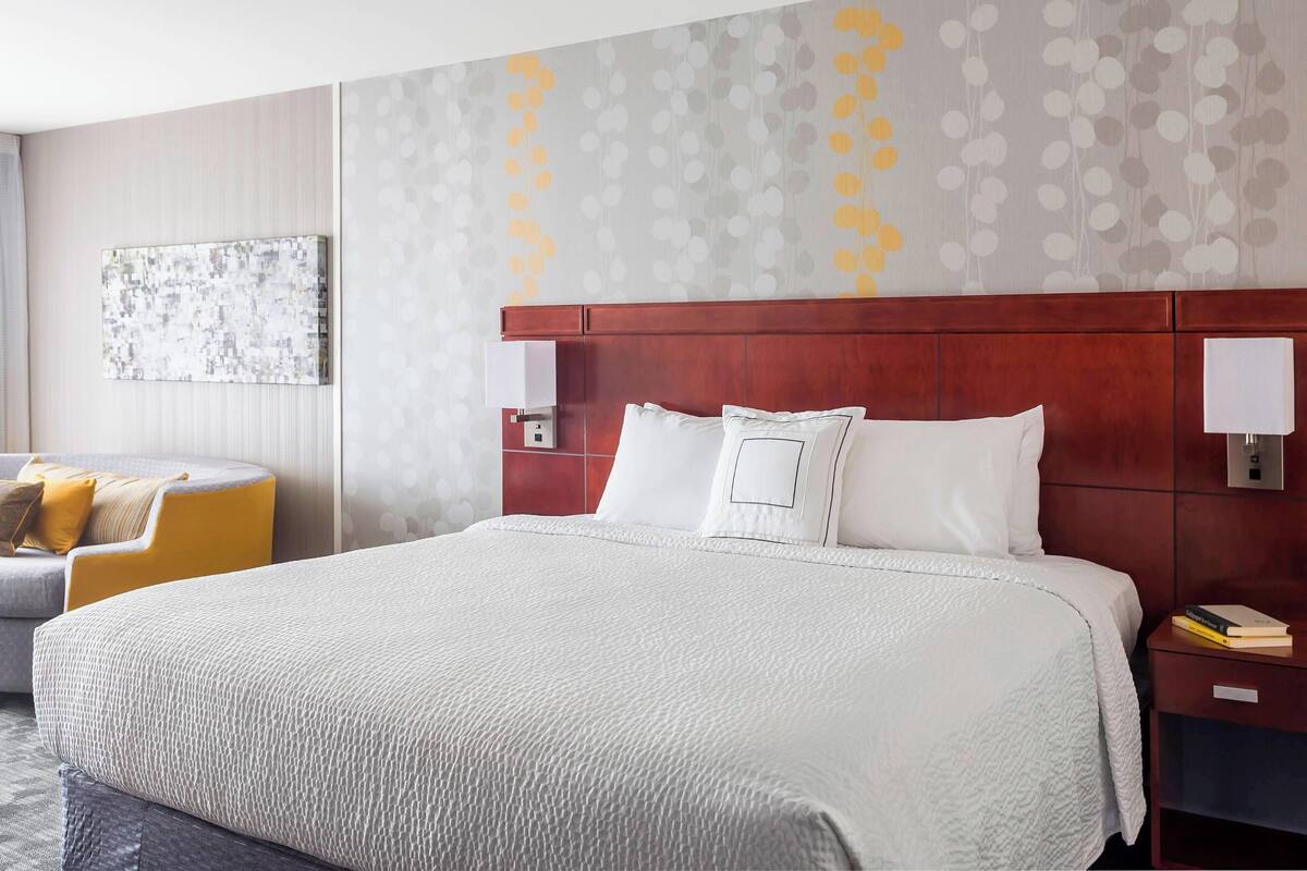 A hotel room with a large bed, a wooden headboard, two bedside tables with lamps, a wall-mounted artwork, and a sofa with cushions.
