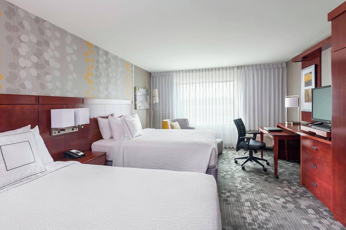 A hotel room with two beds, a desk, a chair, a TV, and a window with curtains.