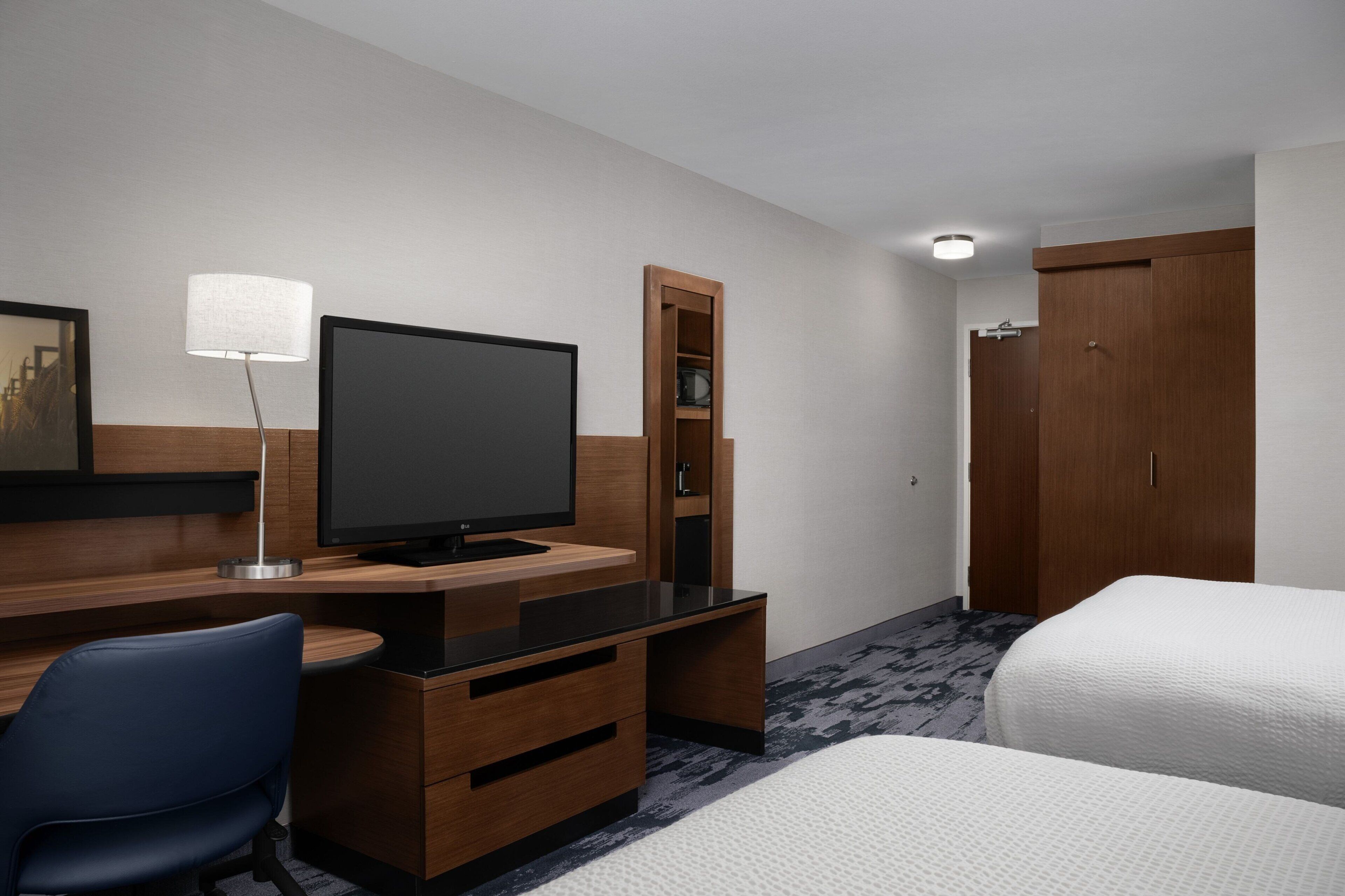 A hotel room with a wooden desk, a television, a blue chair, a bed, and a wardrobe.