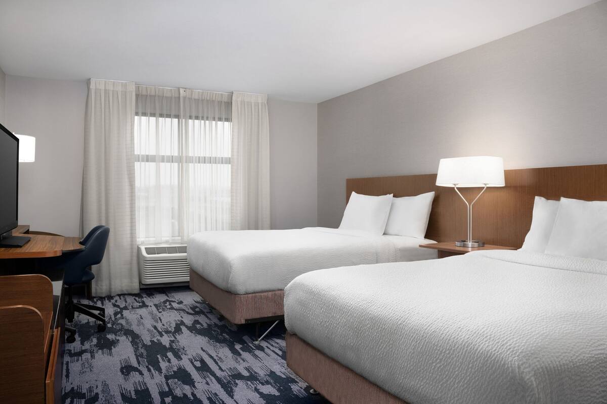 A hotel room with two beds, a desk, a chair, a TV, and a lamp.