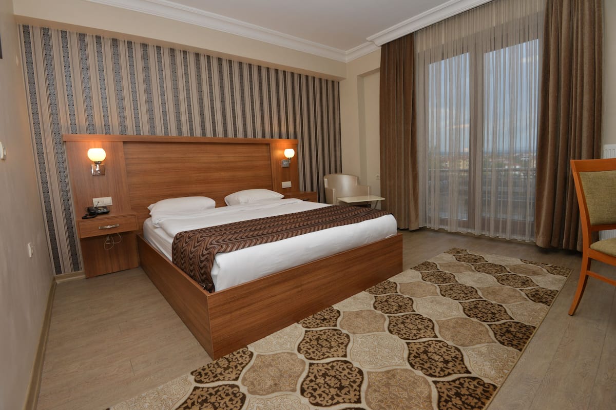 A hotel room with a wooden bed, a nightstand, a lamp, a chair, and a view of the city through large windows.