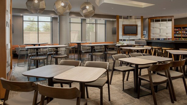 A modern dining area with multiple tables and chairs, large windows, and a television. - SpringHill Suites Jackson North/Ridgeland (Ridgeland)