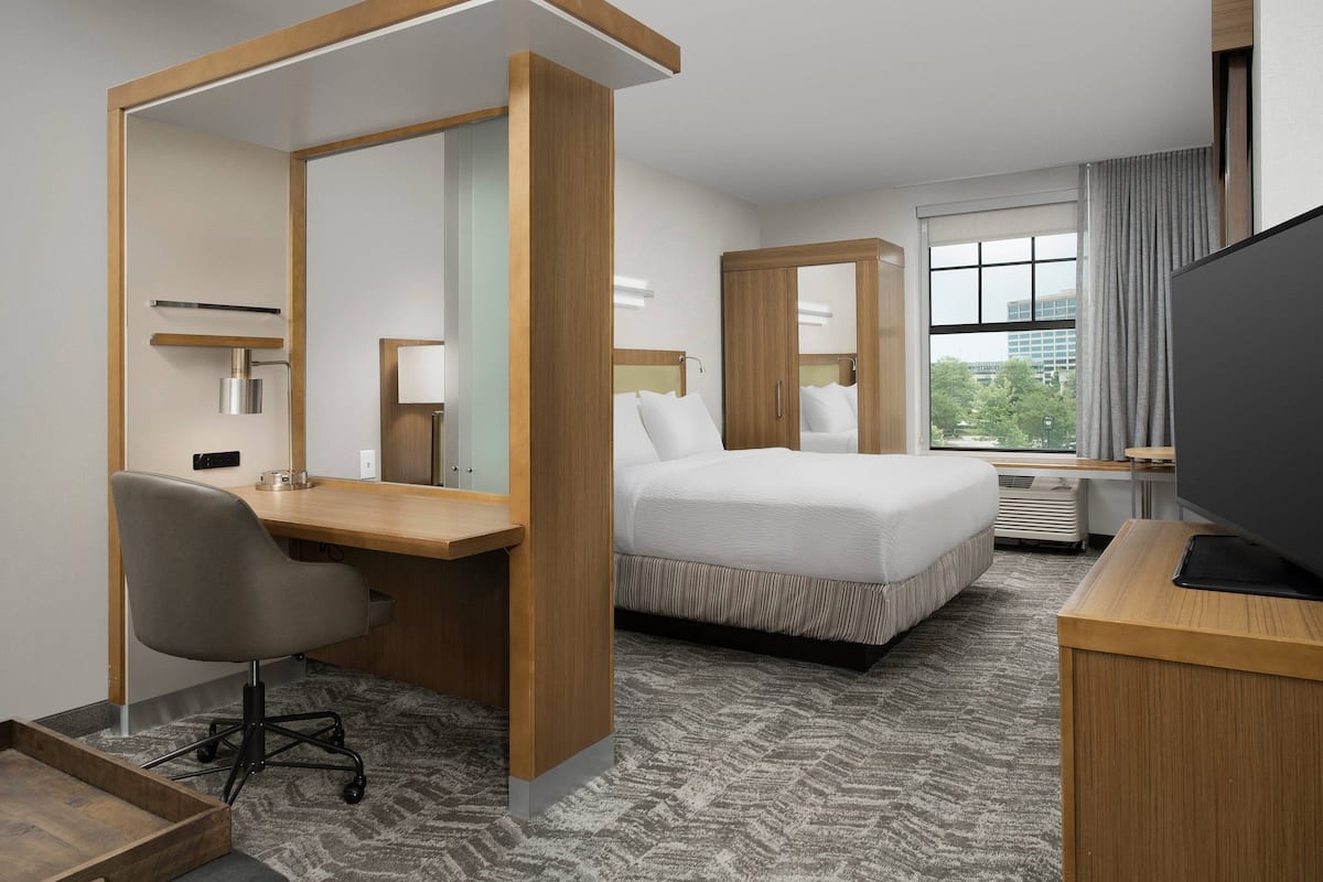 Habitación de hotel con cama, escritorio, silla, televisor y ventanas amplias.