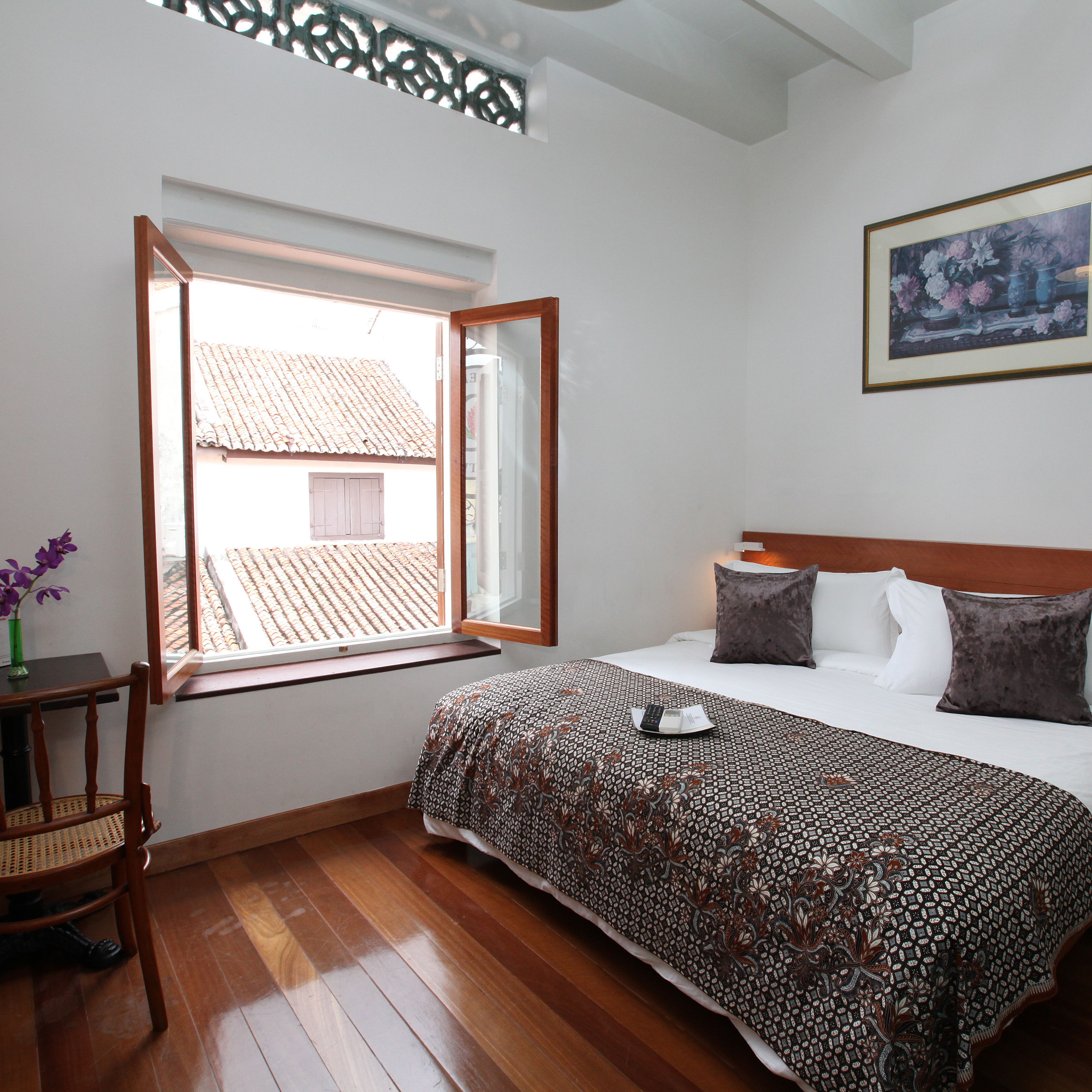A bedroom with a large bed, a window with an open view, a wooden chair, and a framed picture on the wall.