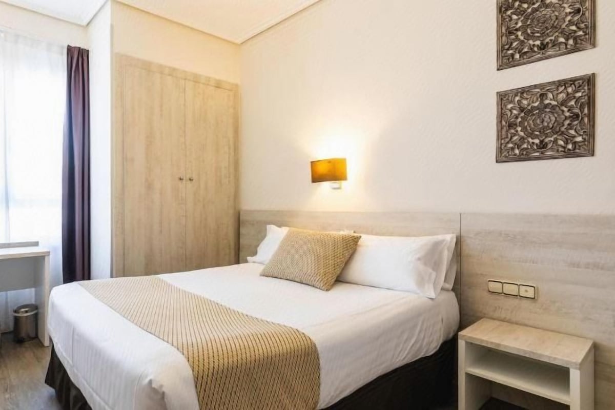 A hotel room with a bed, bedside table, desk, chair, and a window with curtains.