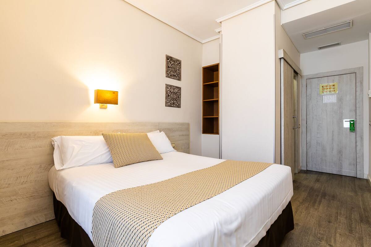 A hotel room with a bed, a wall-mounted light, and a door with a lock.