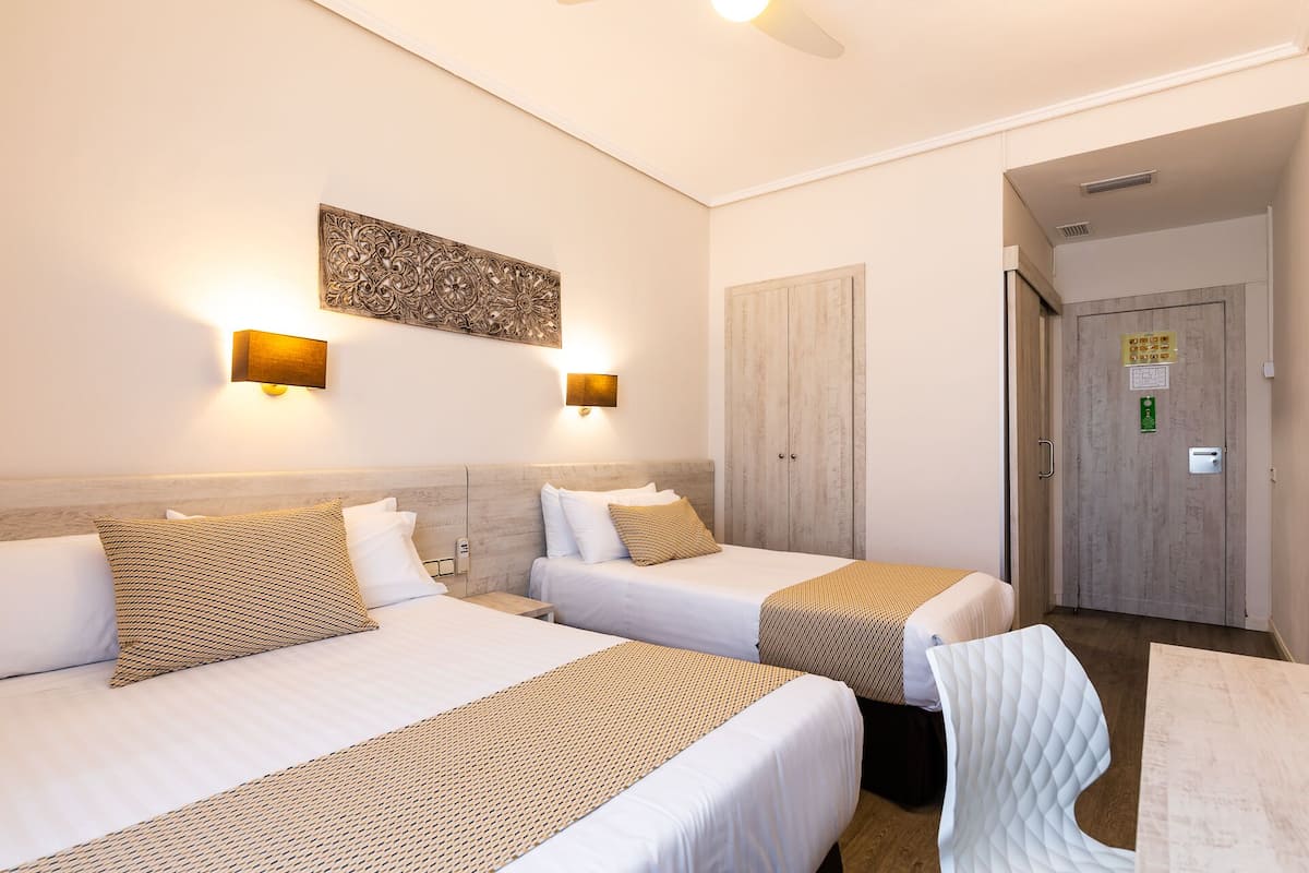 A hotel room with two beds, a wooden headboard, wall-mounted lights, a decorative wall piece, and a door with a sign.