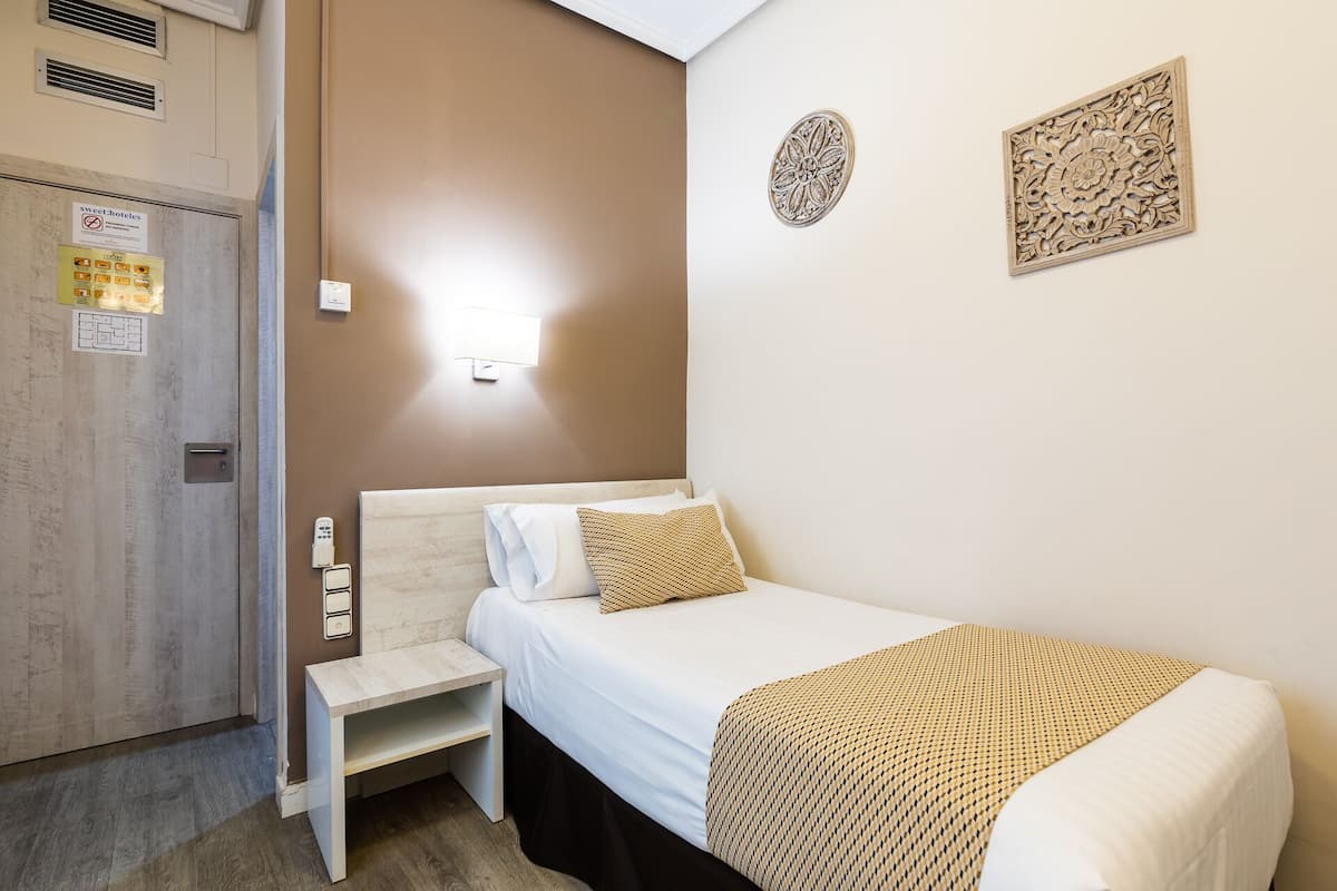 A small, neatly arranged bedroom with a single bed, a bedside table, and a wall-mounted light.