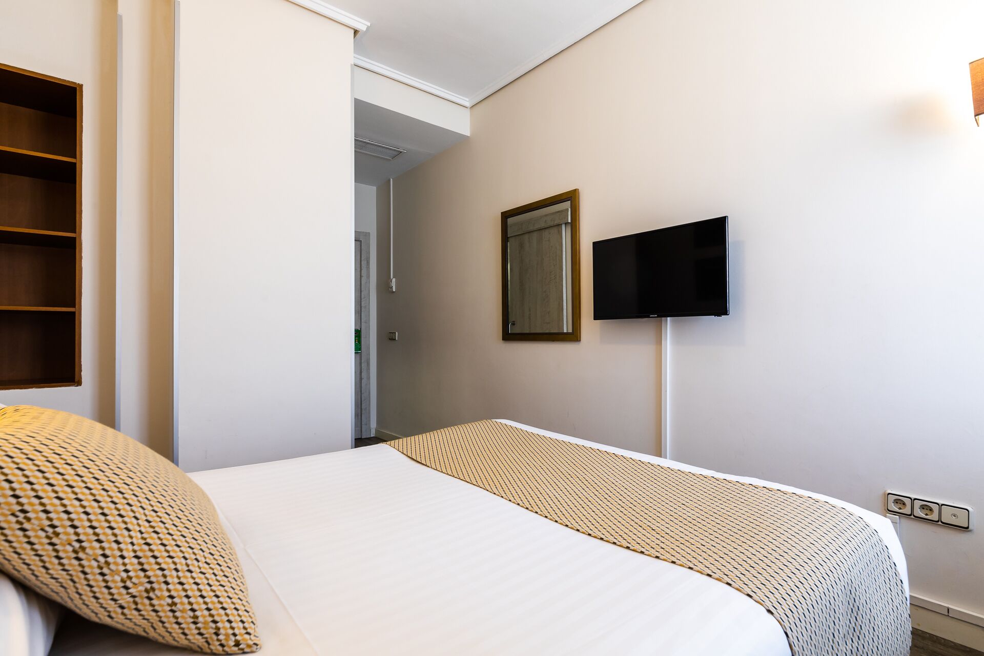 A hotel room with a bed, a television mounted on the wall, and a door leading to a hallway.