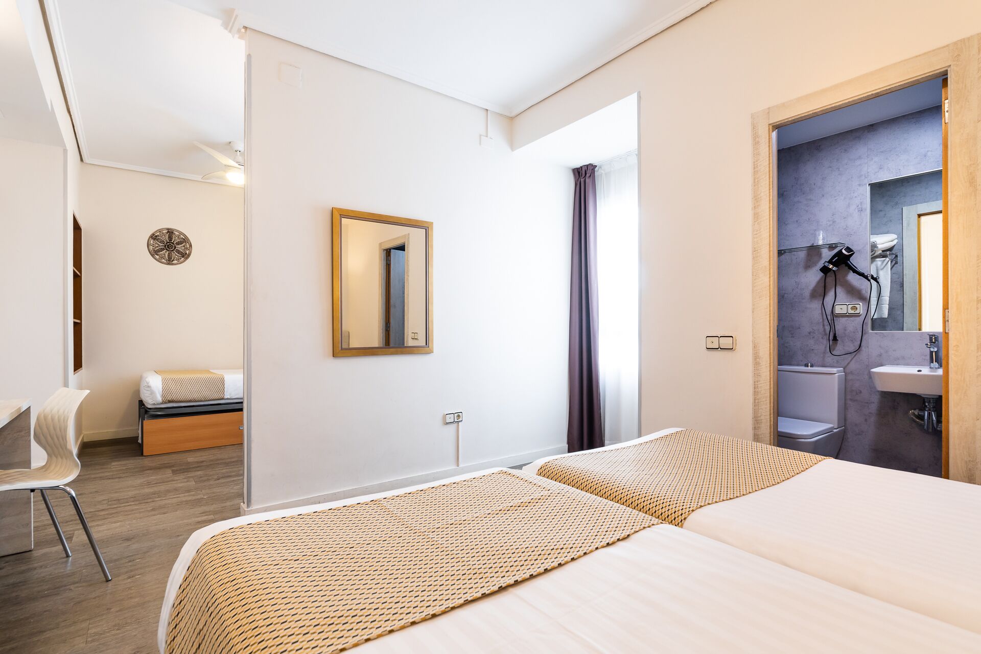 Habitació d'hotel amb dos llits, una taula de treball, una cadira, un mirall i un bany visible per una porta oberta.