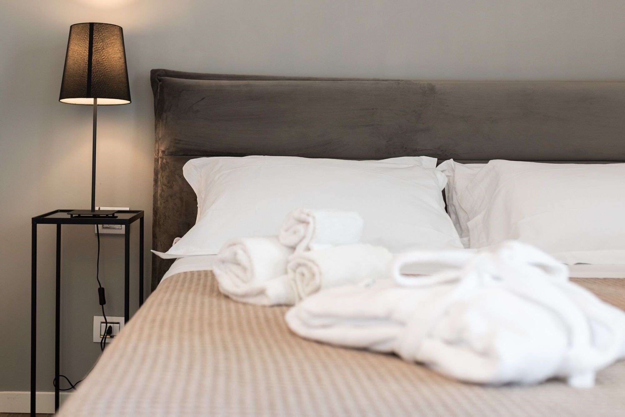 Ein ordentlich bezogenes Bett mit weißen Leinentüchern und aufgerollten Handtüchern, eine moderne Lampe und eine Steckdose auf einem Nachttisch.