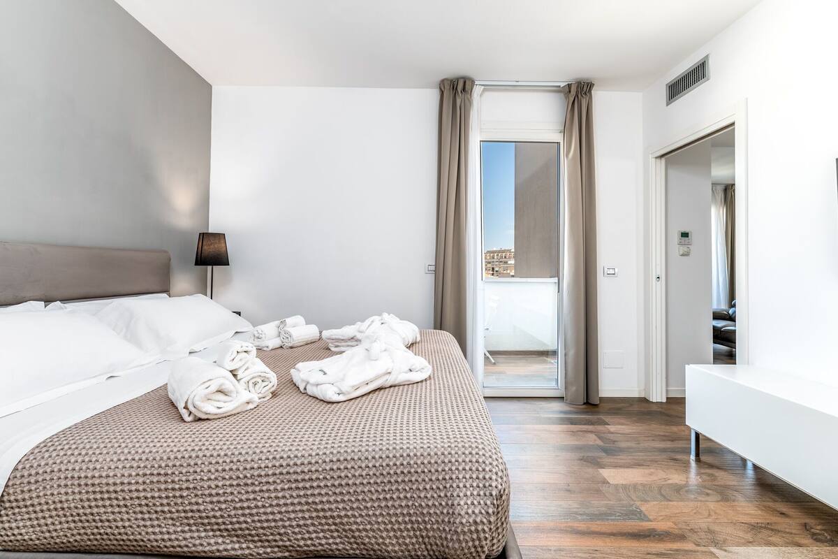 A modern hotel room with a large bed, a bedside lamp, and a view of the city through a glass door.