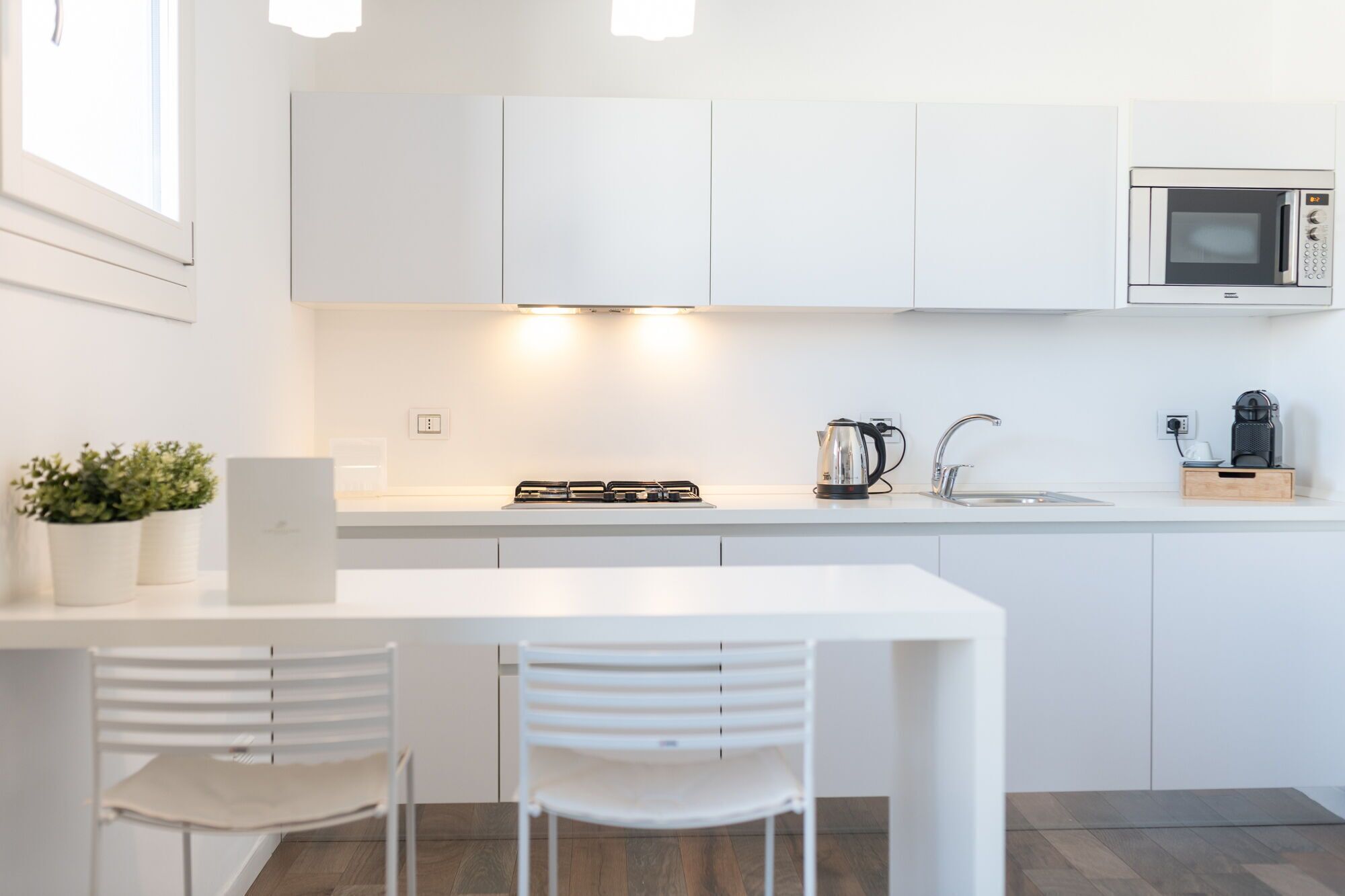 Una cocina moderna con armarios blancos, estufa a gas, hervidor, microondas y un lavamanos. Hay dos sillas blancas en una mesa.
