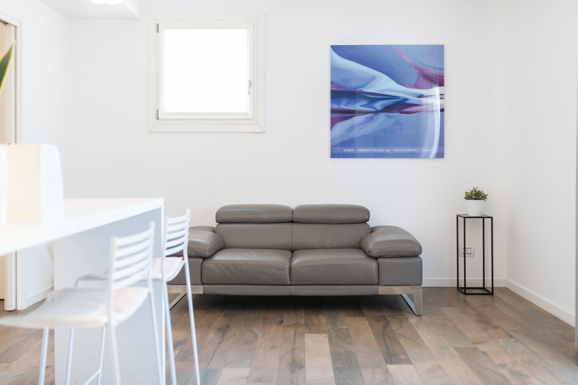 Un salon moderne avec un canapé gris, une petite table d’appoint ornée d’une plante et une fenêtre avec des stores.