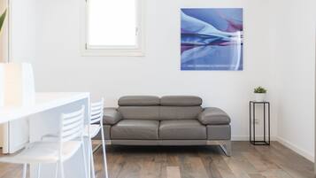 A modern living room with a grey sofa, a small side table with a plant, and a window with blinds.