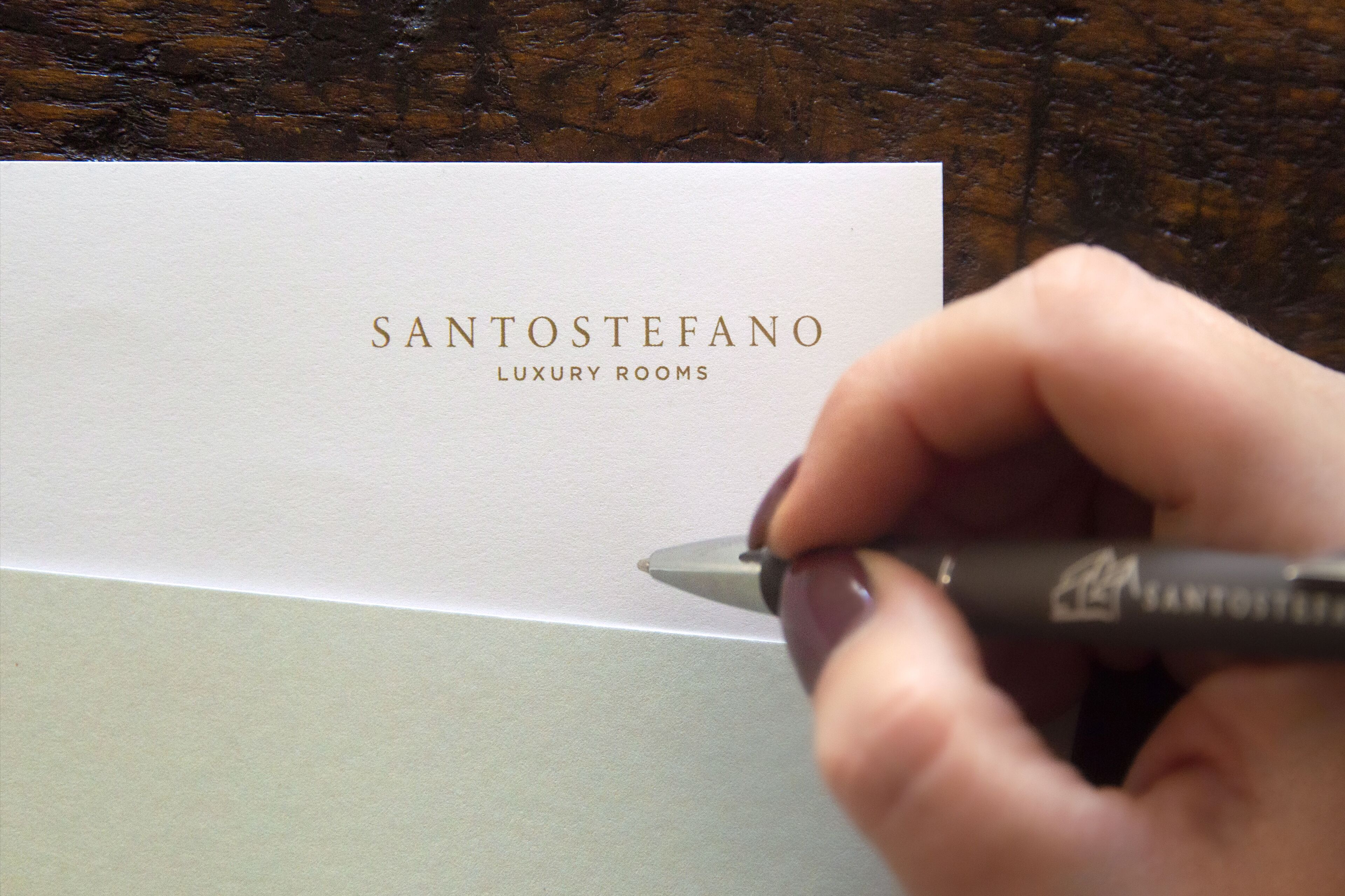 Une main écrivant sur une carte blanche le texte : « SANTOSTEFANO, CHAMBRES DE LUXE ».
