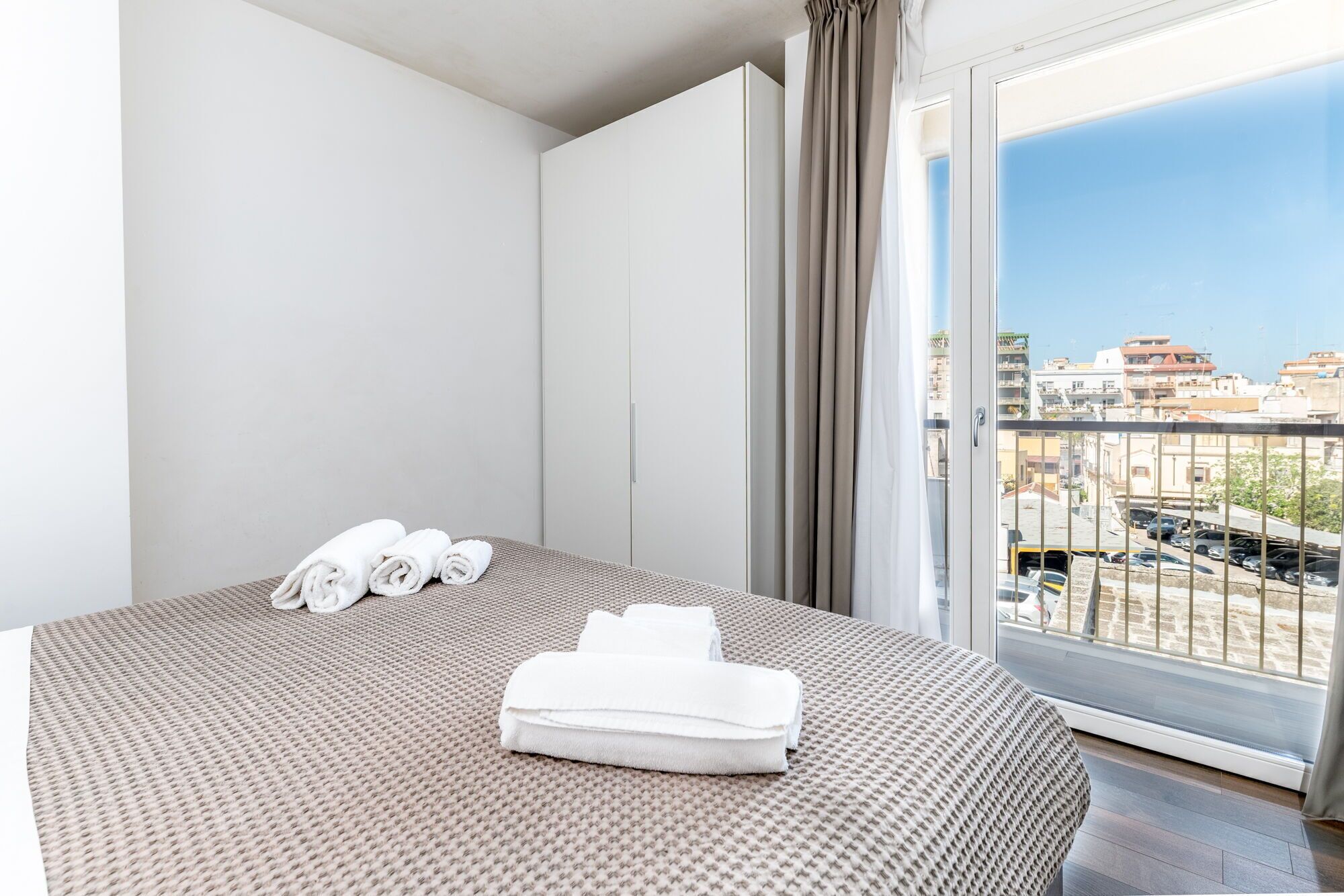 A hotel room with a bed, a balcony with a view of buildings, and a neatly folded towel on the bed.