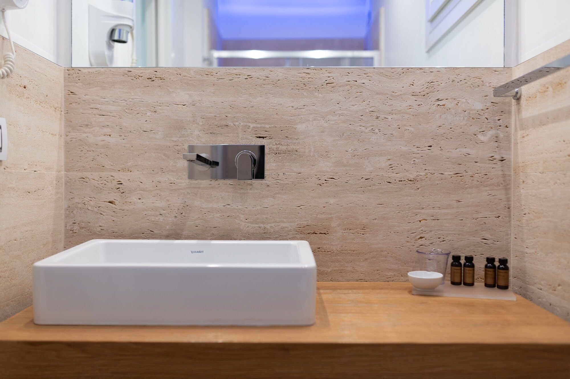 A modern bathroom with a rectangular white bathtub, a wall-mounted faucet, and a wooden vanity with a small bowl and several bottles.