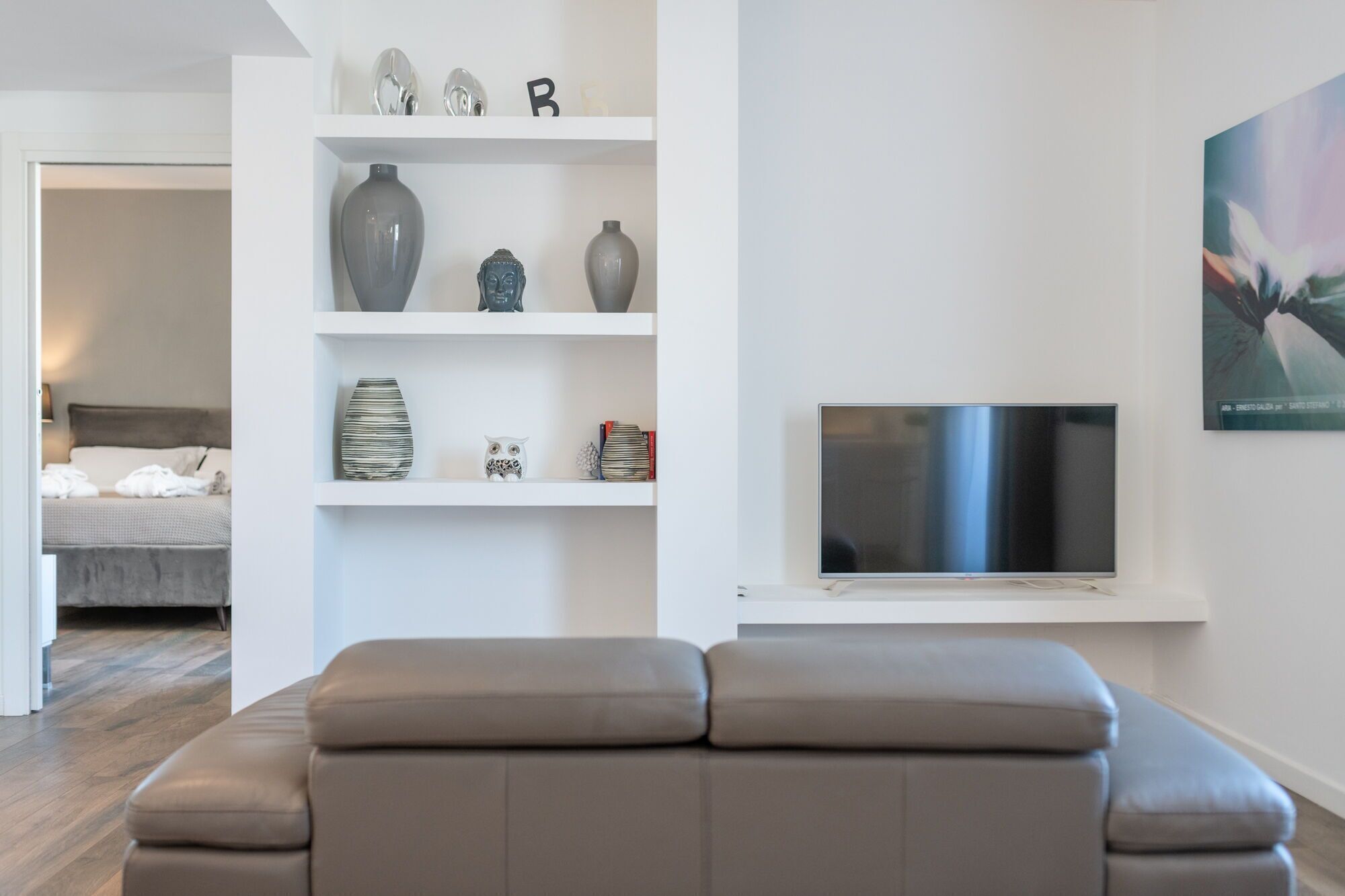 Una sala de estar moderna con un sofá gris, un televisor de pantalla plana y un estante blanco con objetos decorativos.