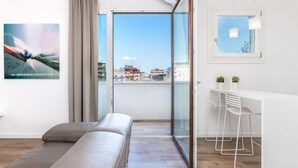 Un salon moderne avec un canapé, un coin repas comprenant une table et des chaises blanches, et une grande porte-fenêtre coulissante donnant sur un balcon avec vue sur la ville.