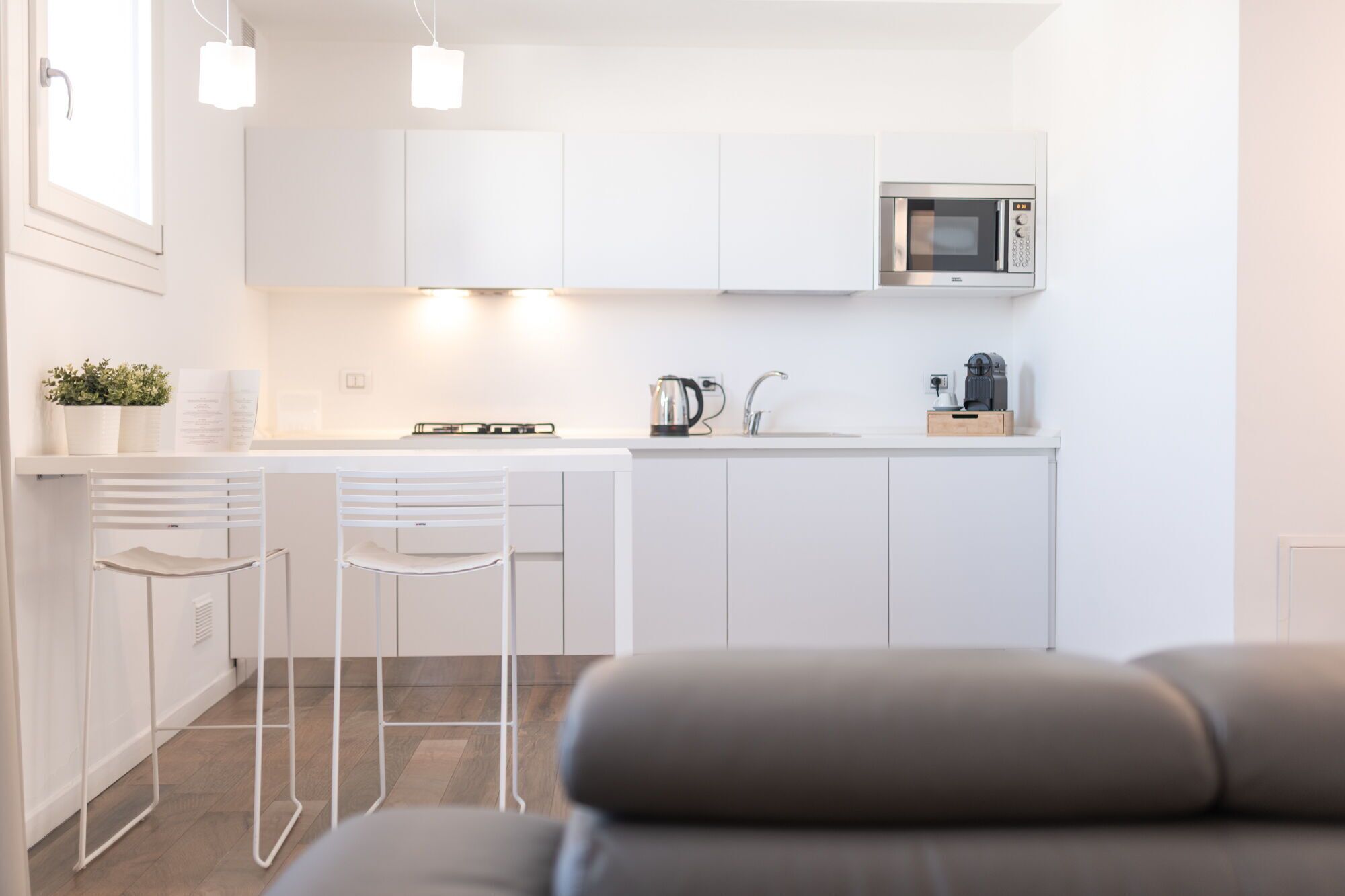 Een moderne keuken met witte kasten, een ingebouwde magnetron, een waterkoker en een koffieapparaat. Op de voorgrond staan twee witte barkrukken en een grijze bank.