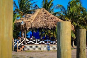 A thatched-roof structure with a white lattice fence, surrounded by palm trees. - Hotel Costa Azul Faro Marejada (Puerto Cortés)