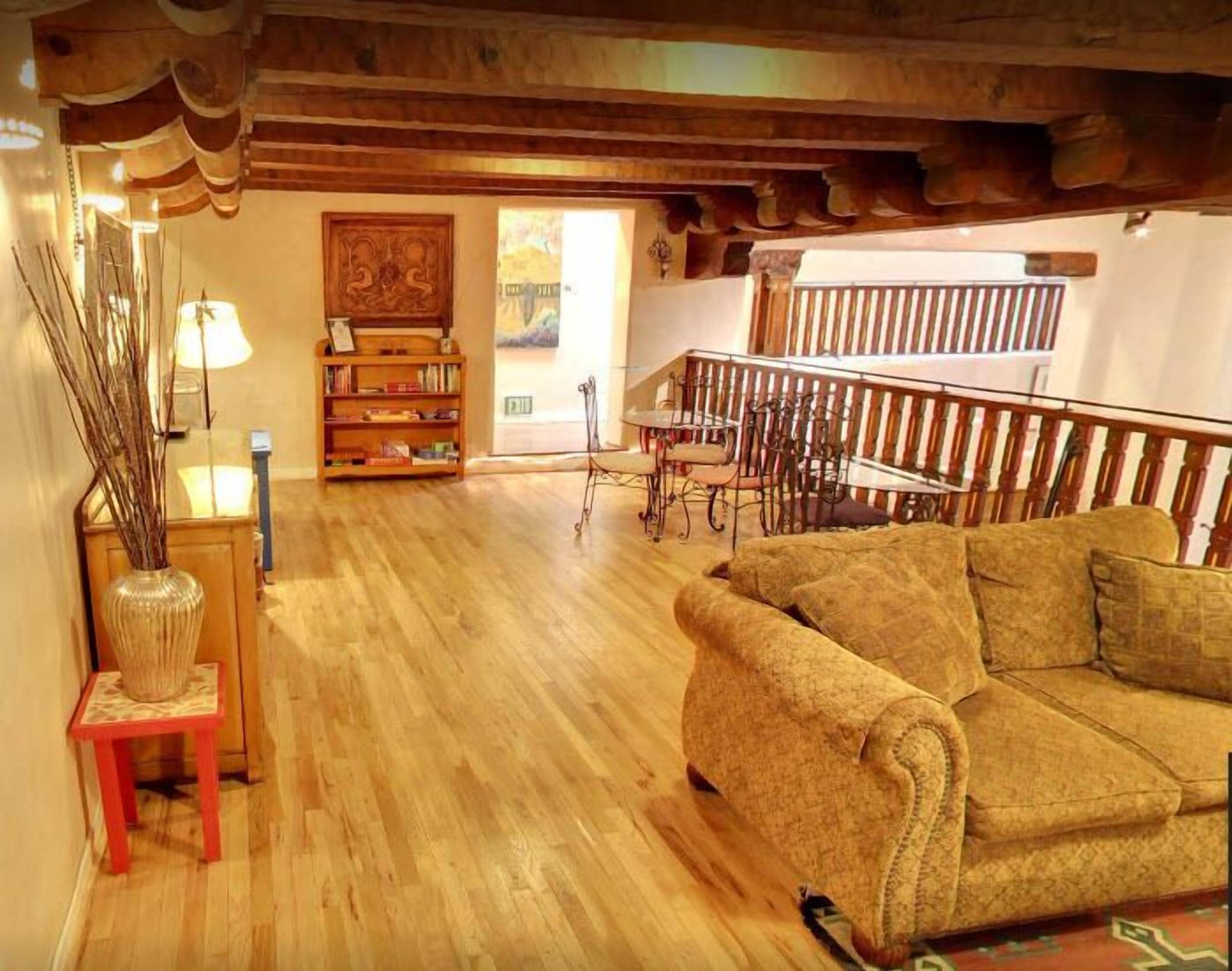 A cozy living room with a wooden floor, a sofa, a bookshelf, and a dining area with a railing.