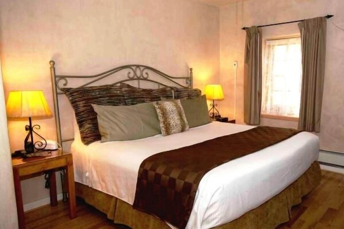 A neatly made bed with a headboard, two bedside lamps, a window with curtains, and a wooden dresser.
