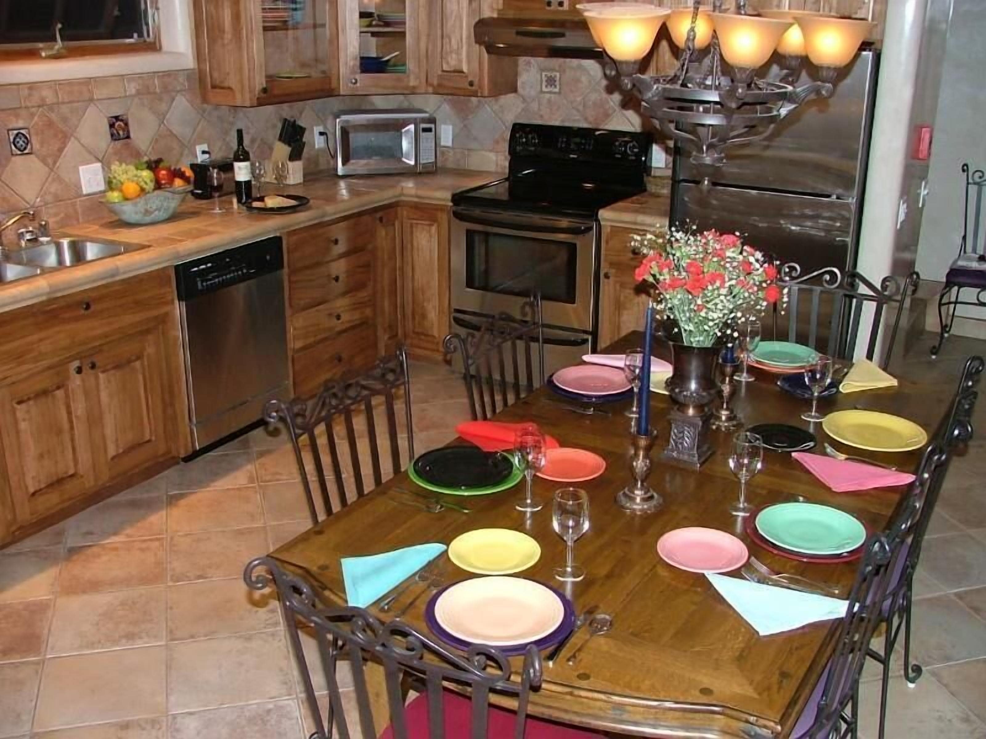 Una mesa de comedor de madera dispuesta con platos coloridos y copas de vino, rodeada de sillas de metal.
