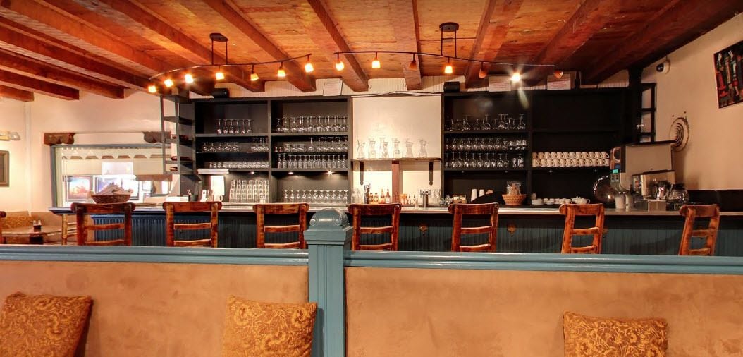 A bar area with wooden chairs and a counter, shelves with glasses and bottles, and a rustic ceiling.