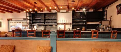 A bar area with wooden chairs and a counter, shelves with glasses and bottles, and a rustic ceiling.
