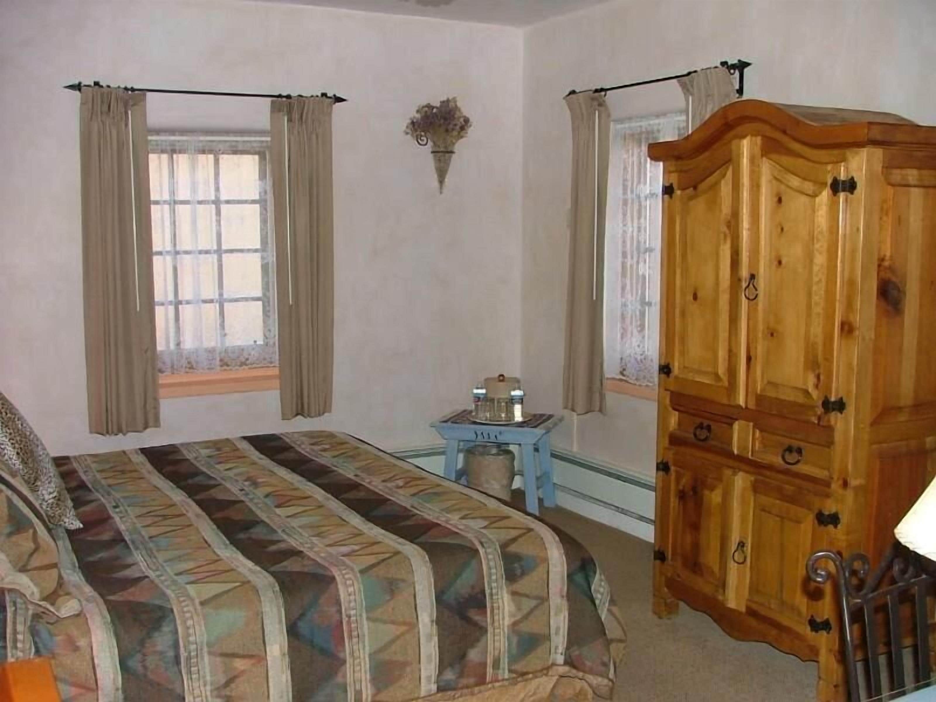 A bedroom with a bed, a wooden wardrobe, a small table with items, and curtains on the windows.