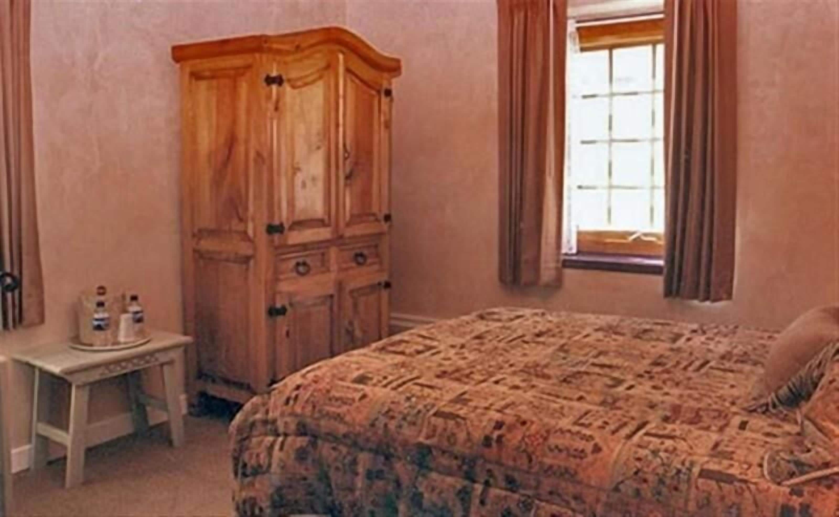 A bedroom with a large wooden wardrobe, a small bedside table with bottles, and a patterned bedspread.