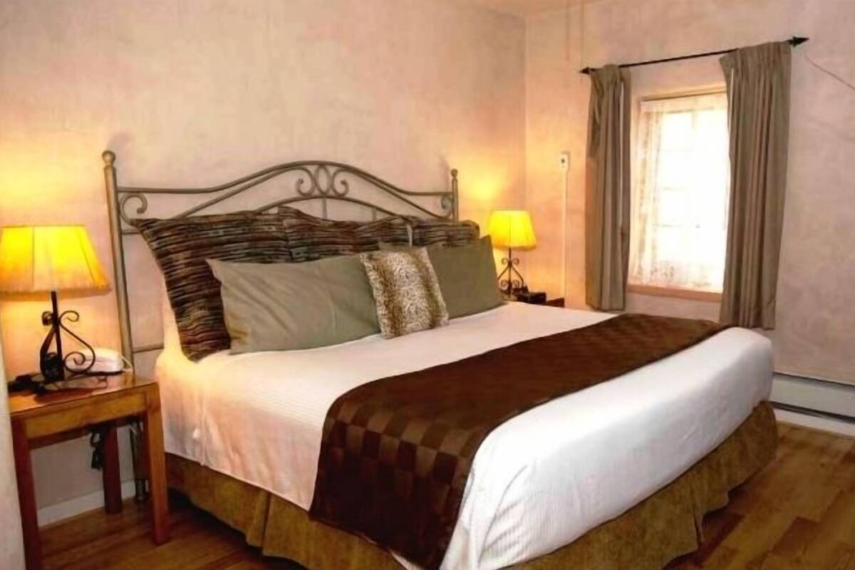 Un letto rifatto con un poggiatesta, due lampade ai lati, una finestra con tende e un comò in legno.