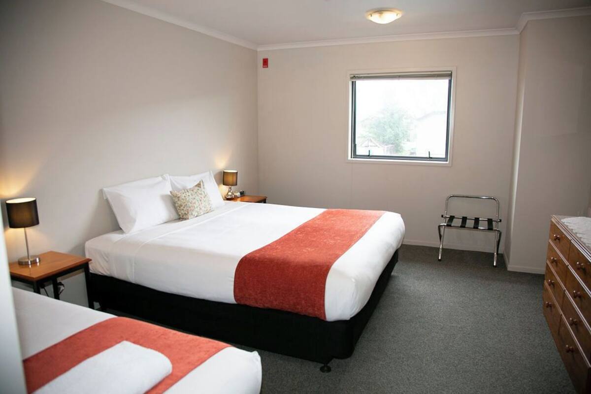 A hotel room with two beds, a window, a dresser, and a small stool.
