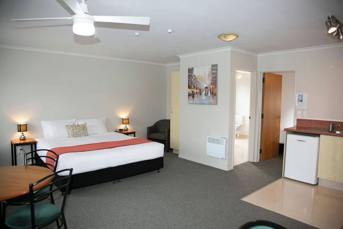 A hotel room with a bed, bedside tables, a desk, a chair, a ceiling fan, a kitchenette, and a door leading to another room.