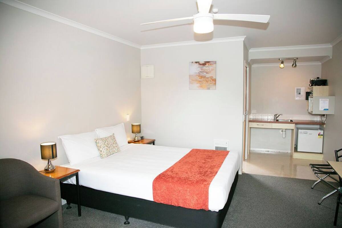 A hotel room with a bed, bedside lamps, a ceiling fan, a small desk, and a kitchenette.