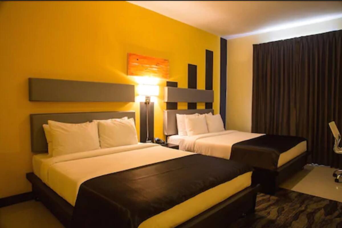 A hotel room with two beds, a desk, a lamp, and a large window with curtains.