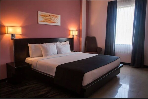 Una habitación de hotel con una cama grande, dos mesitas de noche, una silla y una ventana con cortinas.