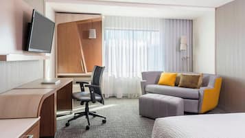 Une chambre d’hôtel équipée d’un bureau, d’une chaise, d’un canapé avec des coussins, d’une télévision et d’une fenêtre avec des rideaux.