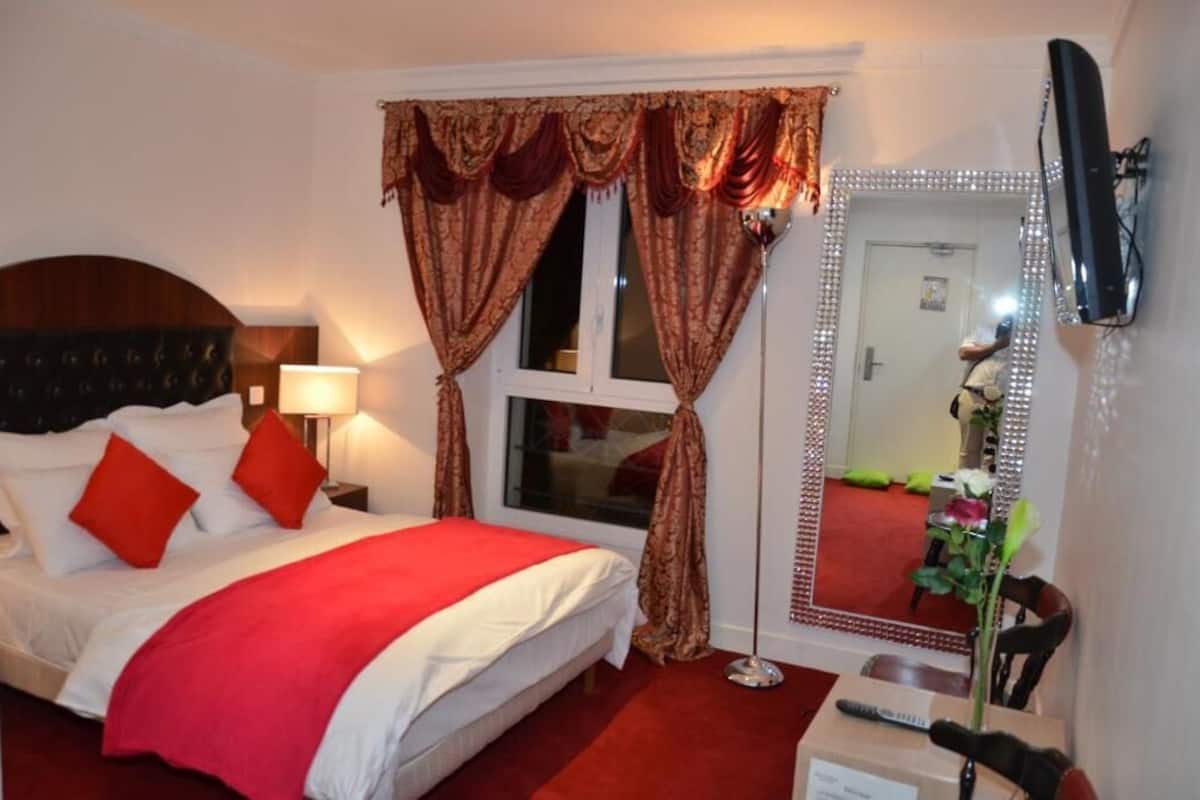 A hotel room with a bed, a bedside table, a lamp, a mirror, and a window with curtains.