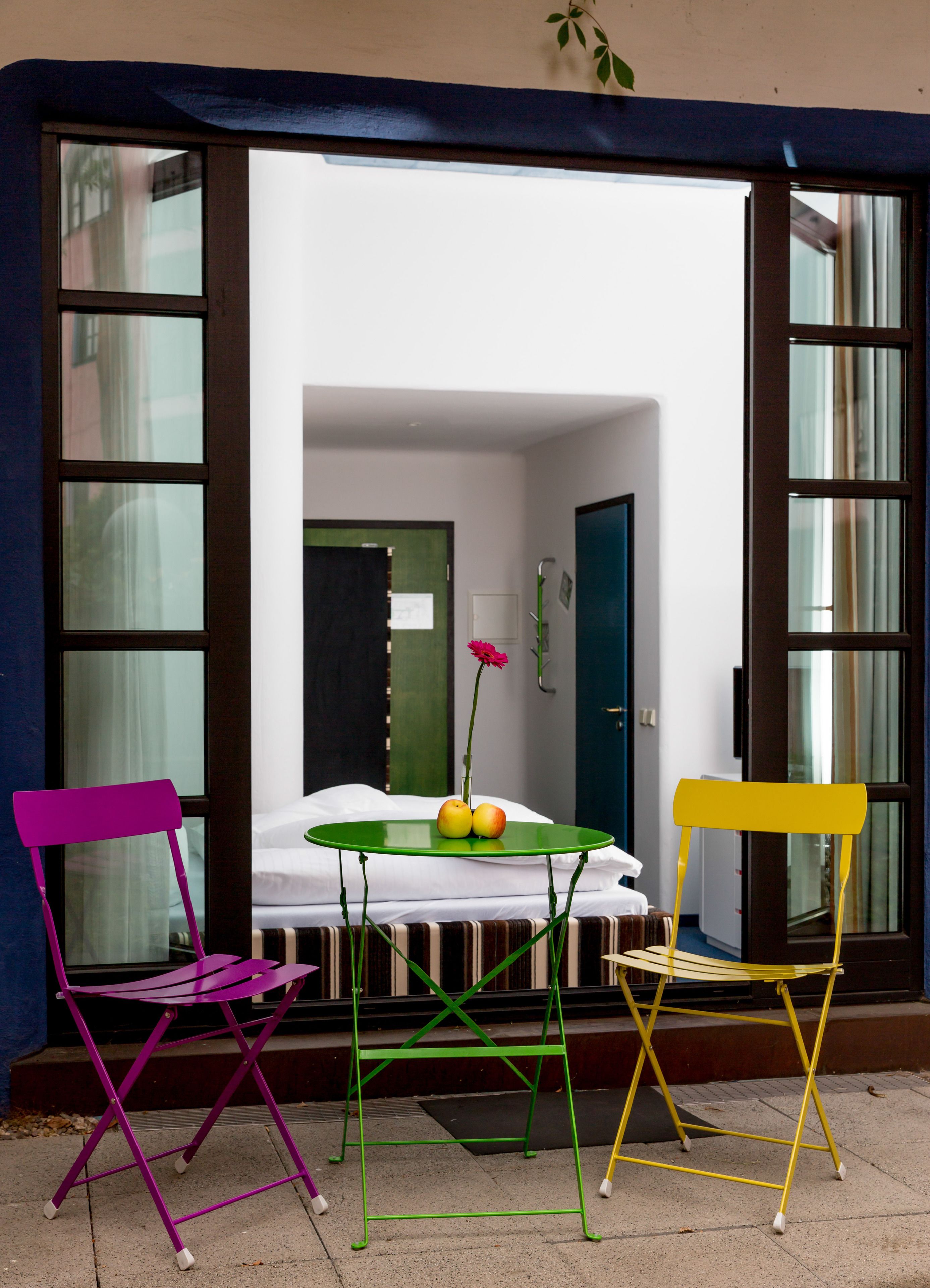 Un pequeño espacio al aire libre con una mesa y dos sillas, un jarrón de flores y vistas a una habitación blanca a través de una puerta de cristal.