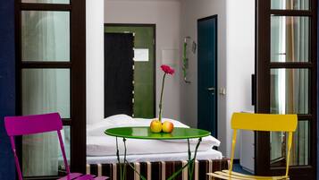 Un pequeño espacio al aire libre con una mesa y dos sillas, un jarrón de flores y vistas a una habitación blanca a través de una puerta de cristal.