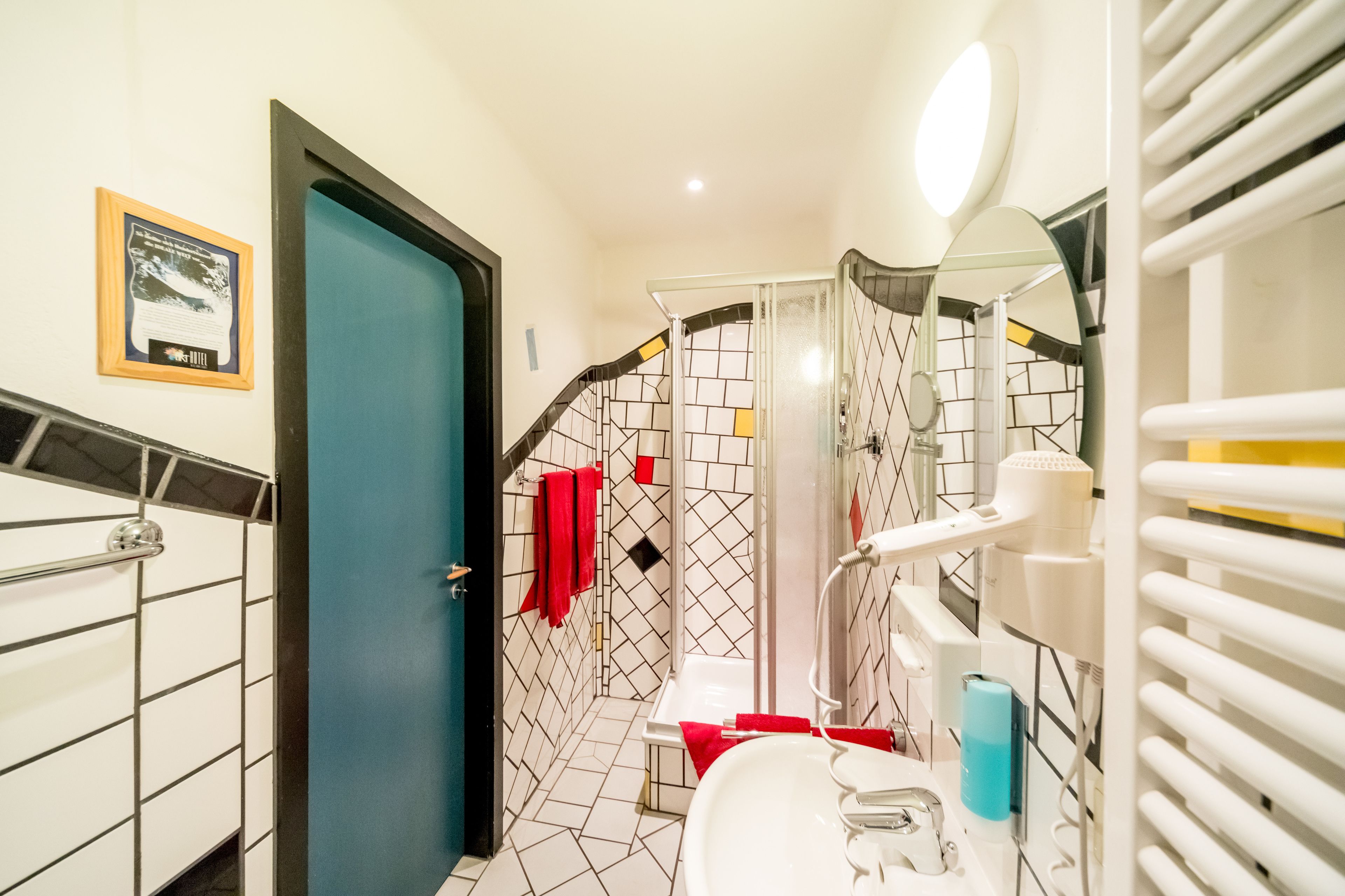 A bathroom with a shower, a sink, and a toilet. There is a towel rack with red towels and a framed picture on the wall.